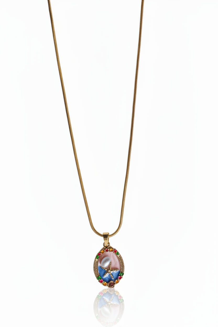 A delicate gold chain necklace featuring an oval pendant with a pink and blue floral design, accented by small multicolored gemstones, shown against a white background with a soft reflection below.