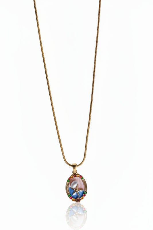 A delicate gold chain necklace featuring an oval pendant with a pink and blue floral design, accented by small multicolored gemstones, shown against a white background with a soft reflection below.